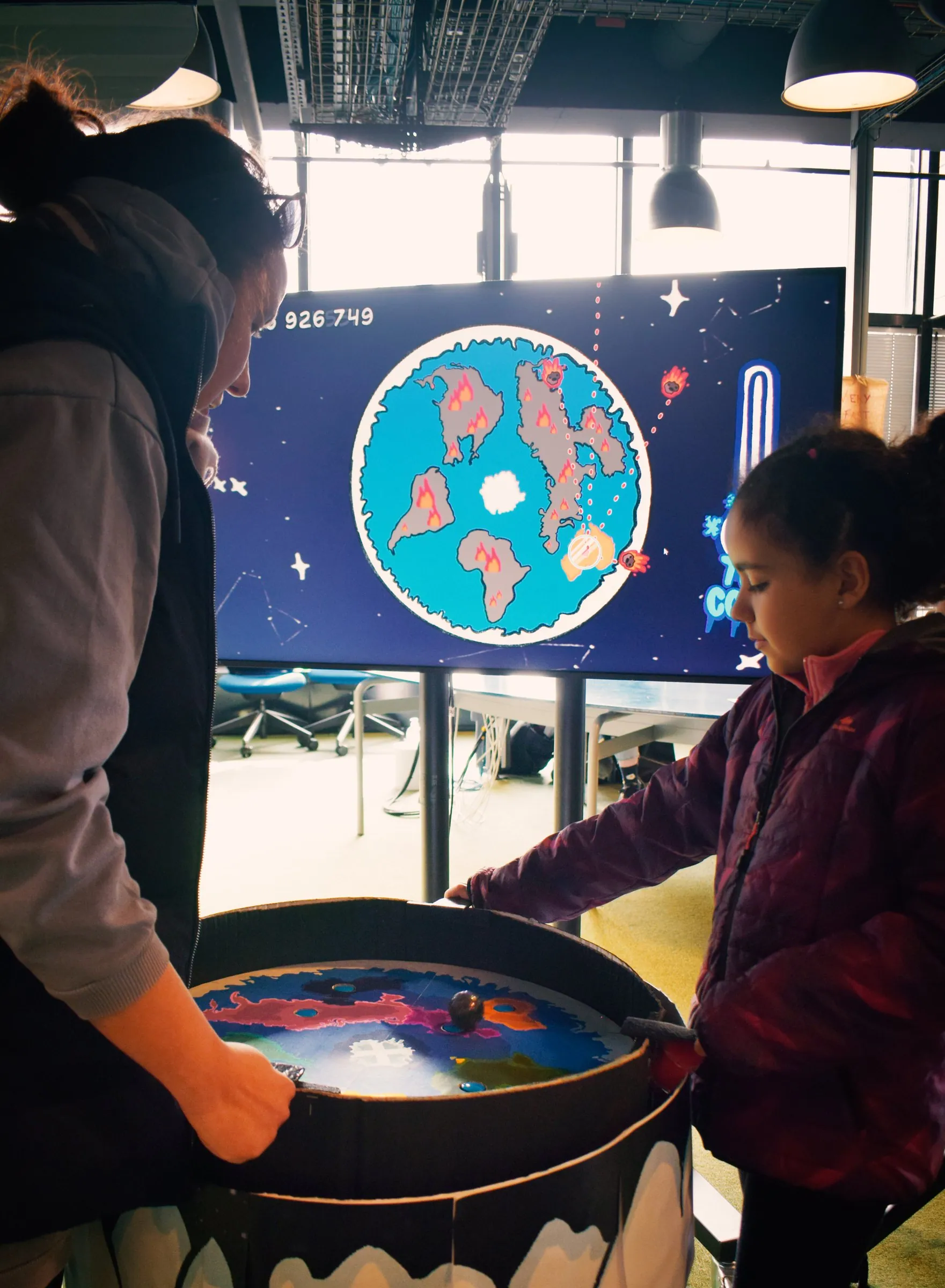 An adult and a child tilting the table, the game on a screen behind them.