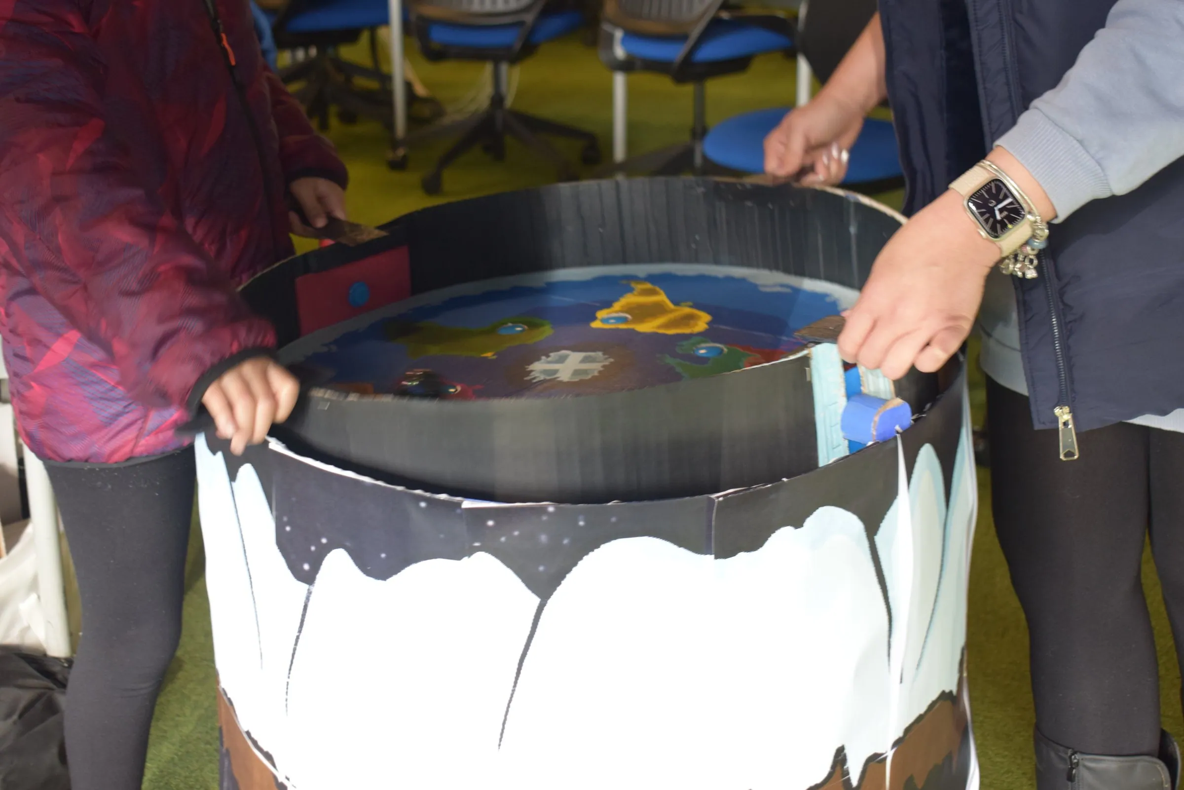 Several hands tilting a round painted table with a ball on its surface.